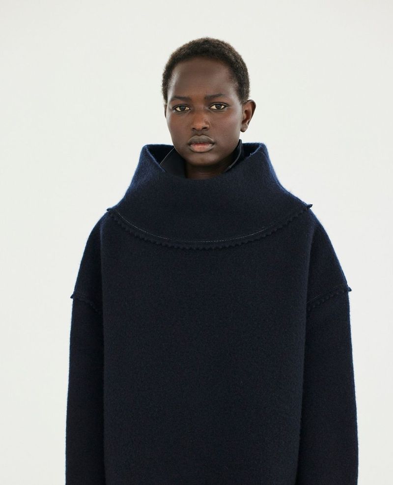 A dark-skinned woman poses indoors wearing a navy blue sweater with a large collar. The photo is a medium shot against a bright background.