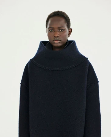 Model Nyaduola Gabriel wearing a dark navy structured wool sweater with a high cowl neckline against a white seamless background.