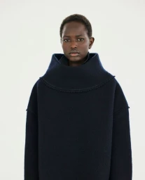 Model Nyaduola Gabriel wearing a dark navy structured wool sweater with a high cowl neckline against a white seamless background.