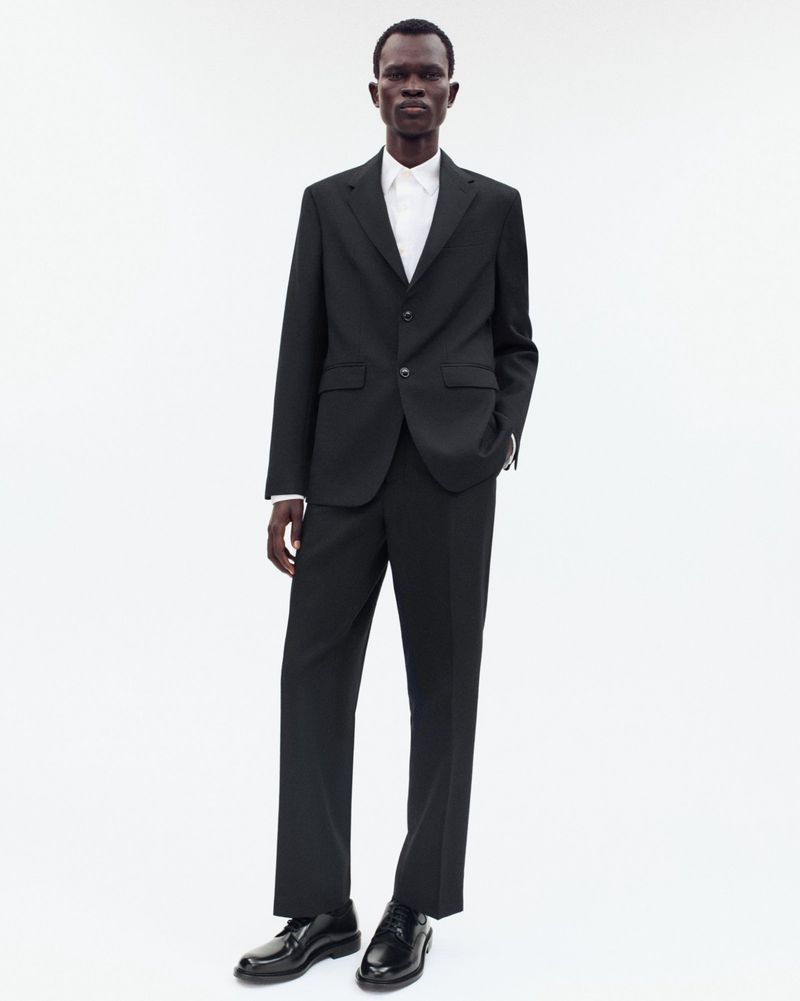 An African-American man in a black suit and white shirt poses against a white backdrop in a fashion-forward studio portrait.