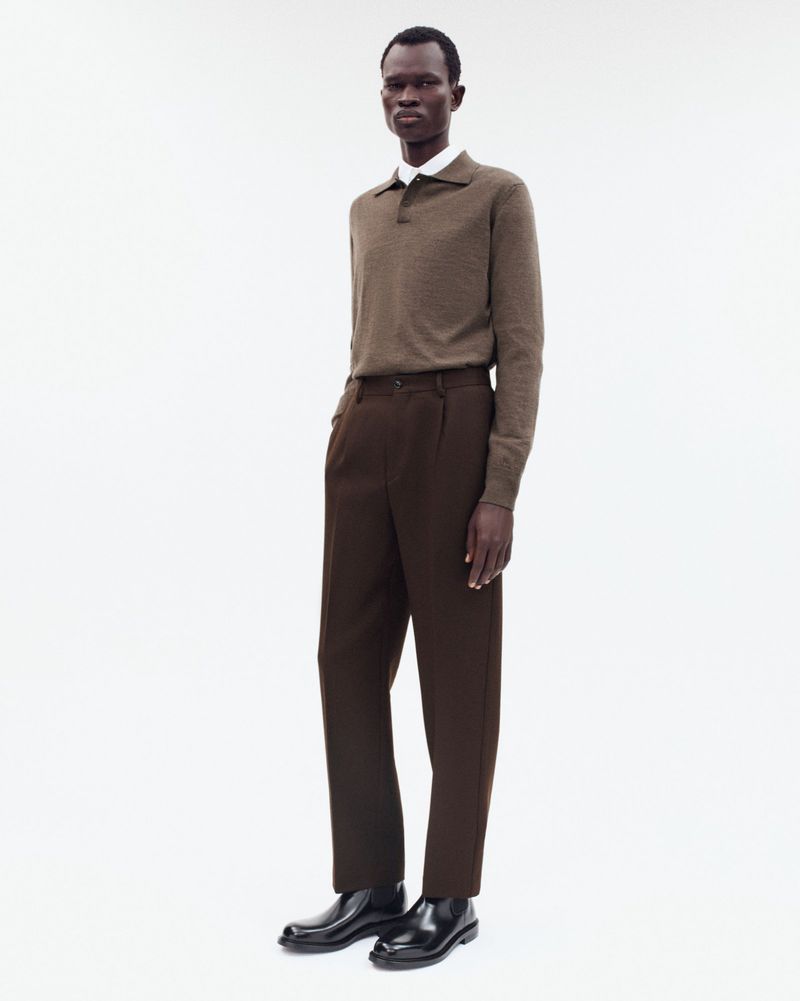 Full-length studio shot of a slender Black male model wearing a brown polo shirt, dark-brown dress pants, and black dress shoes on white.