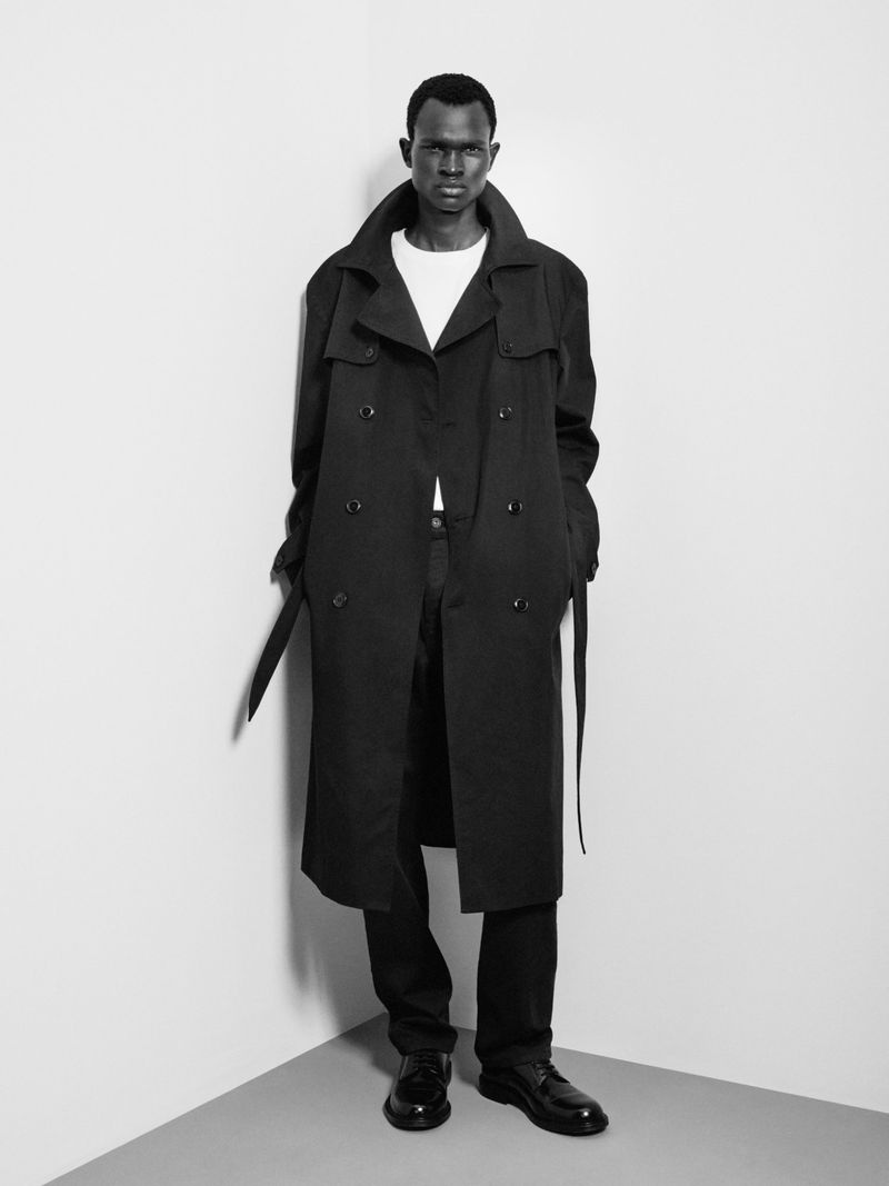 Black and white portrait of a black male model posing in a dark trench coat and black pants, set against a neutral studio background.