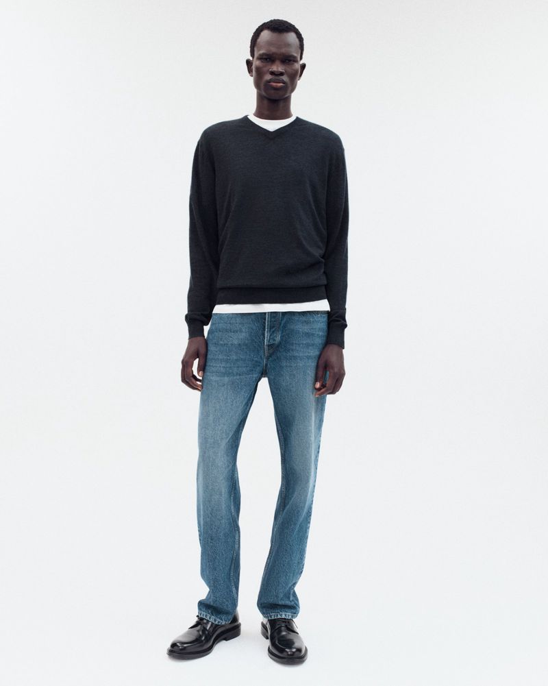 Full-length shot of a dark-skinned male model wearing a black v-neck sweater, jeans, and loafers against a stark white background.