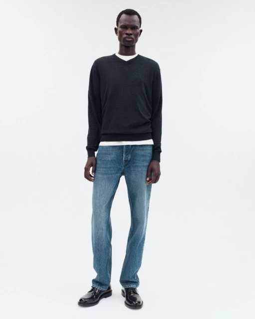 Full-length shot of a dark-skinned male model wearing a black v-neck sweater, jeans, and loafers against a stark white background.