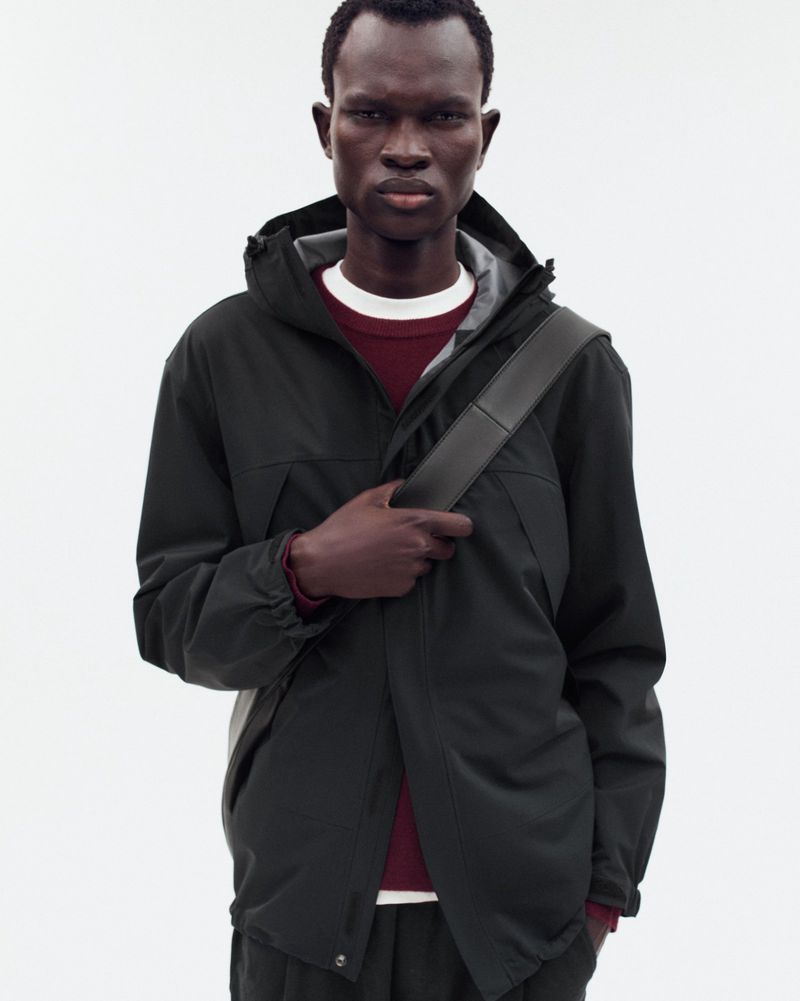 A dark-skinned adult male poses indoors, wearing a hooded green rain jacket, dark pants, and a shoulder bag against a white background.