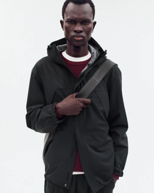 A dark-skinned adult male poses indoors, wearing a hooded green rain jacket, dark pants, and a shoulder bag against a white background.