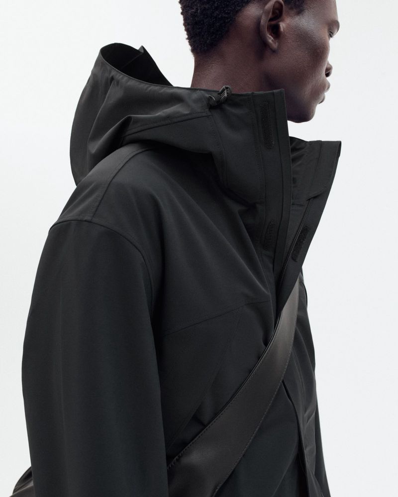 Adult male in a hooded black jacket stands in a neutral studio setting. The camera is at eye level, showcasing fashion and apparel.