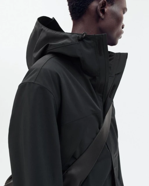 Adult male in a hooded black jacket stands in a neutral studio setting. The camera is at eye level, showcasing fashion and apparel.