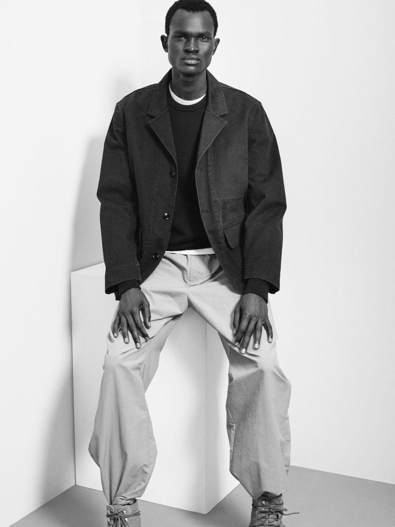 Black and white shot of an adult male in a black jacket, sweater, and tan pants, sitting on a white cube in a studio setting.