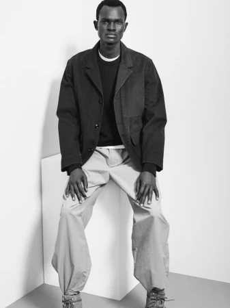 Black and white shot of an adult male in a black jacket, sweater, and tan pants, sitting on a white cube in a studio setting.