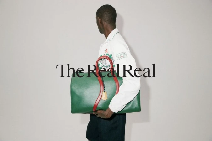 Model holding a large green and red leather duffel bag against a plain light grey studio background for The RealReal
