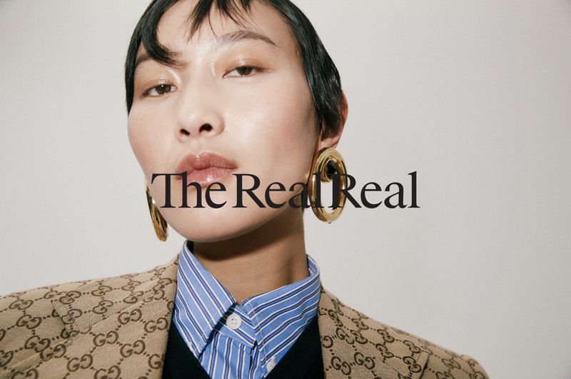 Close-up portrait of a model wearing gold earrings, blue striped shirt, and monogram jacket against a plain background