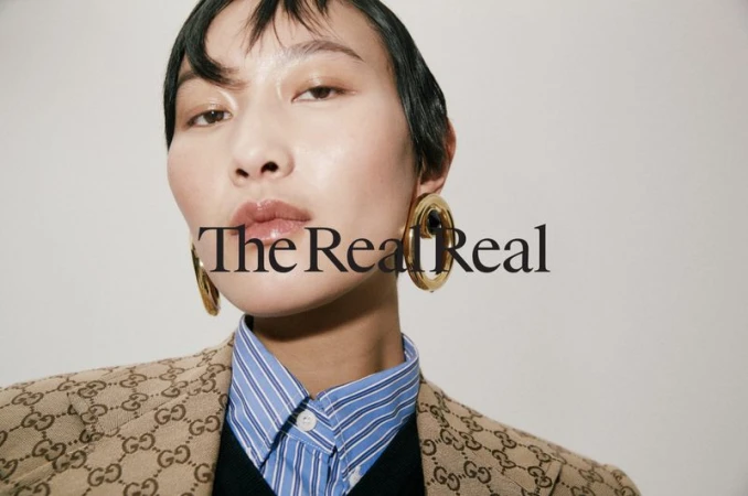 Close-up portrait of a model wearing gold earrings, blue striped shirt, and monogram jacket against a plain background
