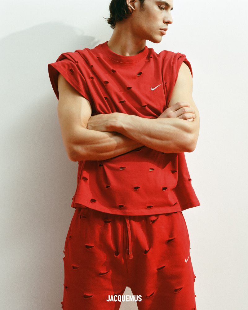 A young male model poses indoors against a light wall, wearing a red, distressed, sleeveless shirt and matching sweatpants with a Nike logo.