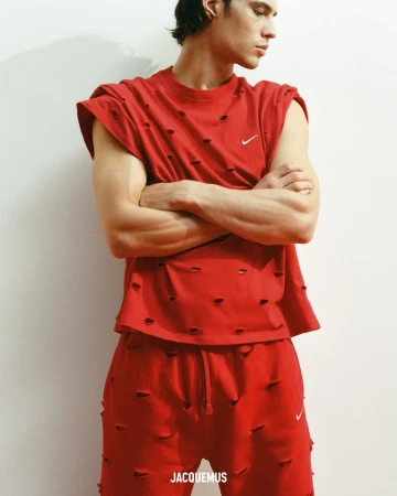 A young male model poses indoors against a light wall, wearing a red, distressed, sleeveless shirt and matching sweatpants with a Nike logo.