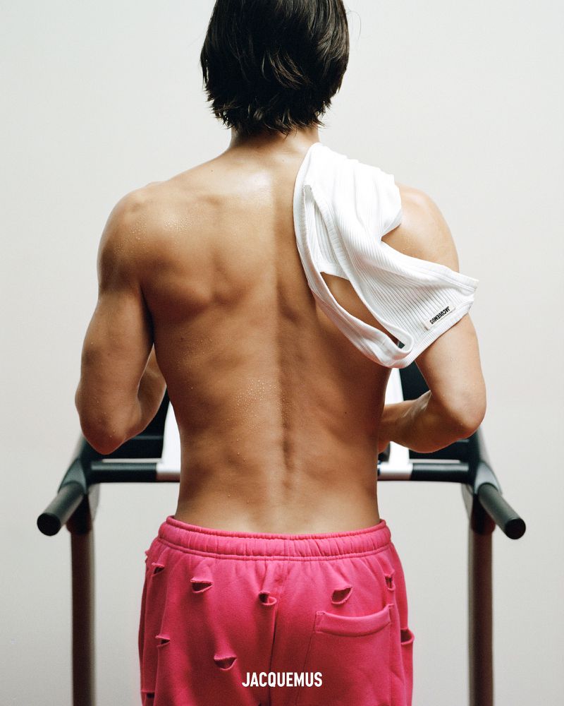 Man on a treadmill with a towel over his shoulder, wearing pink, distressed sweatpants and facing away from the camera against a white wall.