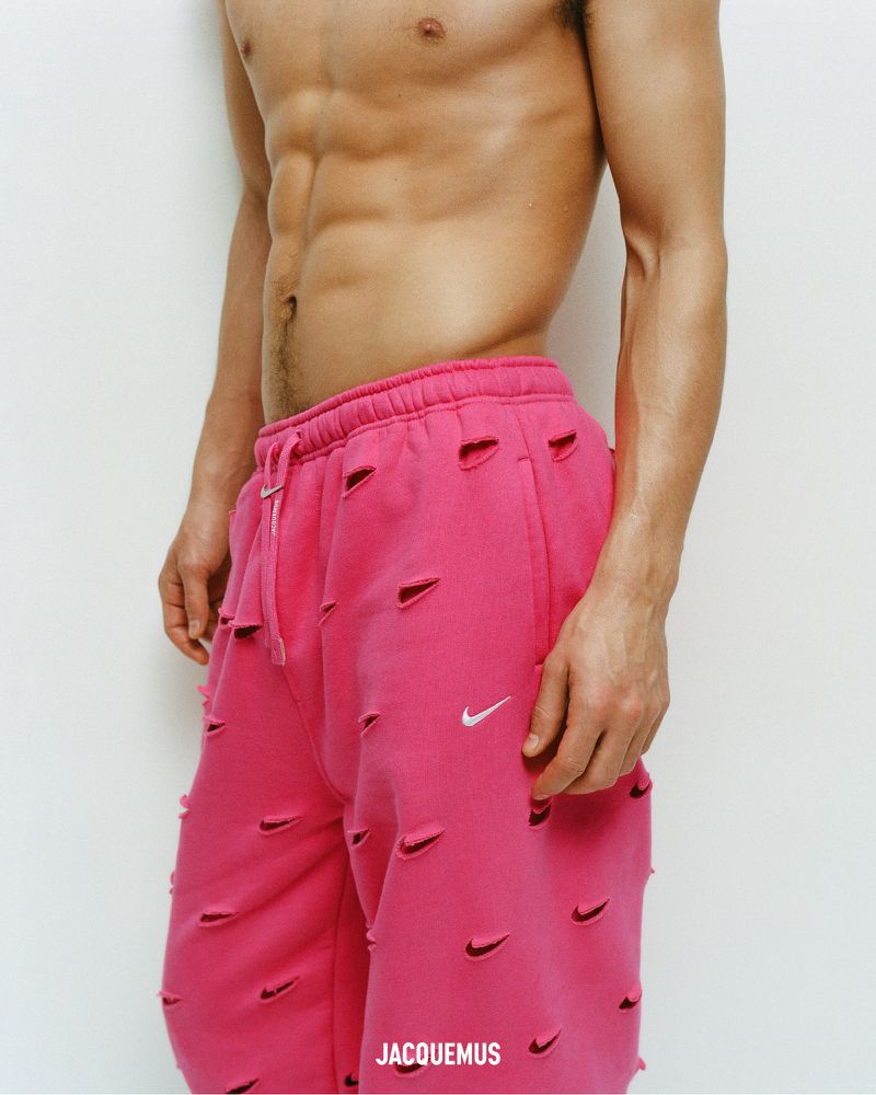 Athletic, shirtless adult man with pink, logo-covered pants poses against a white wall in a studio setting. Fashion, fitness theme.