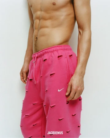 Athletic, shirtless adult man with pink, logo-covered pants poses against a white wall in a studio setting. Fashion, fitness theme.