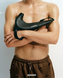 Shirtless male model holding a black leather Jacquemus Nike handbag against a white studio background.