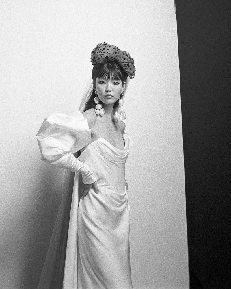 Model in white bridal gown with voluminous sleeves wearing a spotted bow headpiece and pearl earrings.
