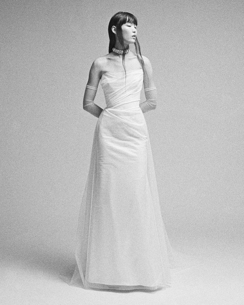 Monochrome portrait of a woman with bangs wearing a white gown and sheer gloves, standing in front of a plain grey backdrop.