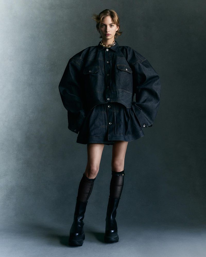 A full-length shot of a woman in a dark denim ensemble, featuring an oversized jacket and mini skirt, complemented by black boots.