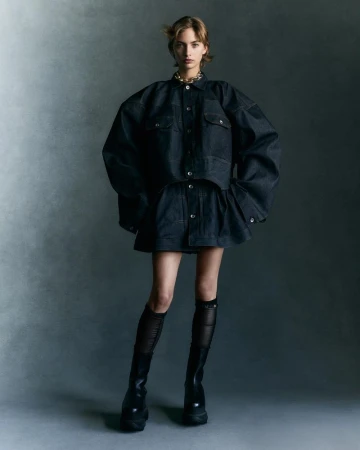 A full-length shot of a woman in a dark denim ensemble, featuring an oversized jacket and mini skirt, complemented by black boots.