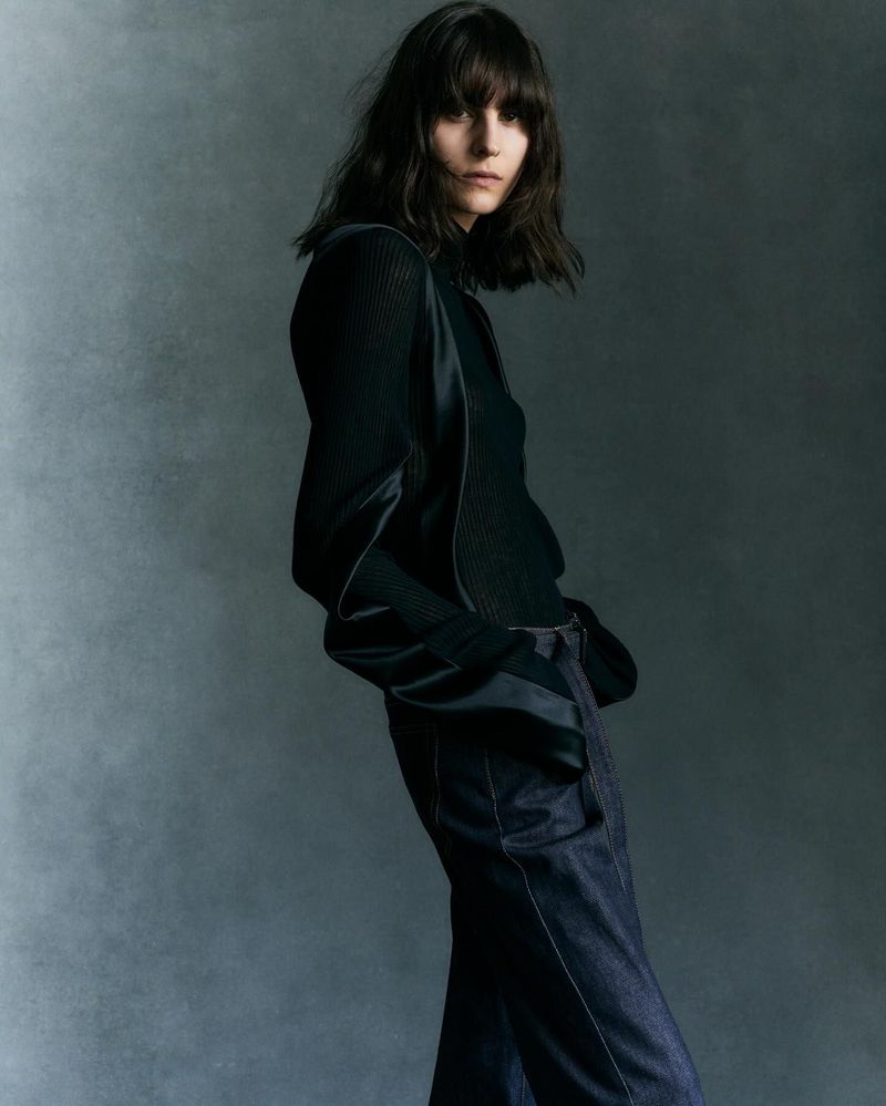 Brunette woman with dark hair in dark blouse and jeans posing in front of gray background, fashion editorial photograph.