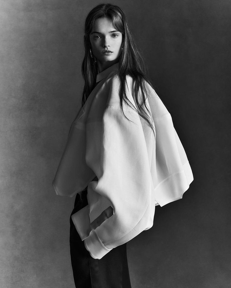 Black and white portrait of a young woman with long hair wearing a white oversized shirt in a studio setting against a grey backdrop.