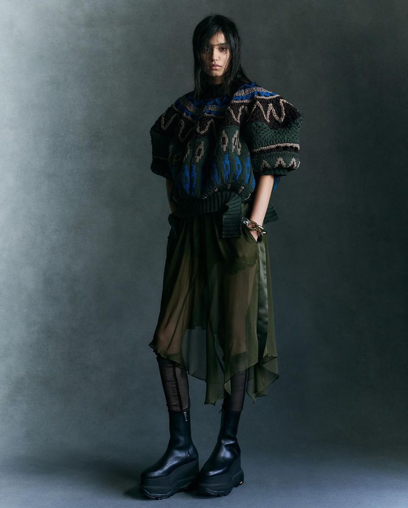 Full-length shot of a woman wearing a patterned knit sweater, sheer layered skirt, platform boots, and dark tights in a studio setting.