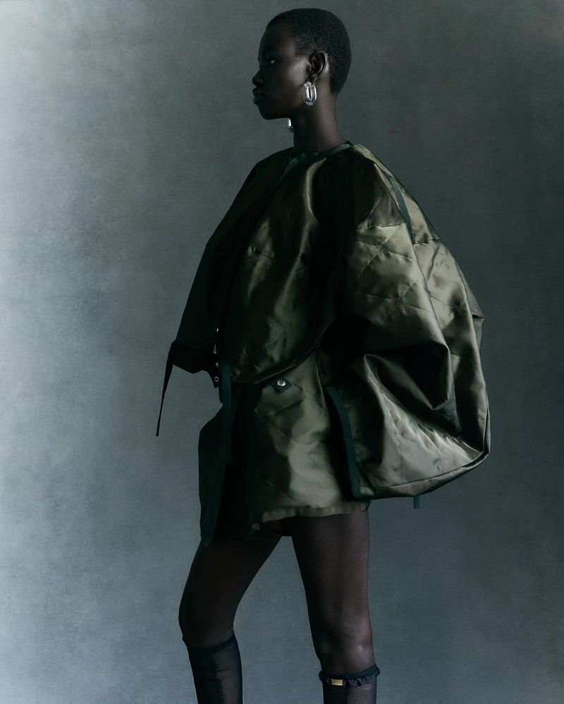 Fashion portrait of an adult woman with dark skin wearing an oversized olive green jacket, sheer black tights, and hoop earrings.
