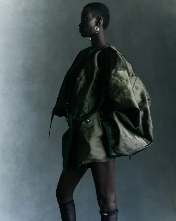 Fashion portrait of an adult woman with dark skin wearing an oversized olive green jacket, sheer black tights, and hoop earrings.