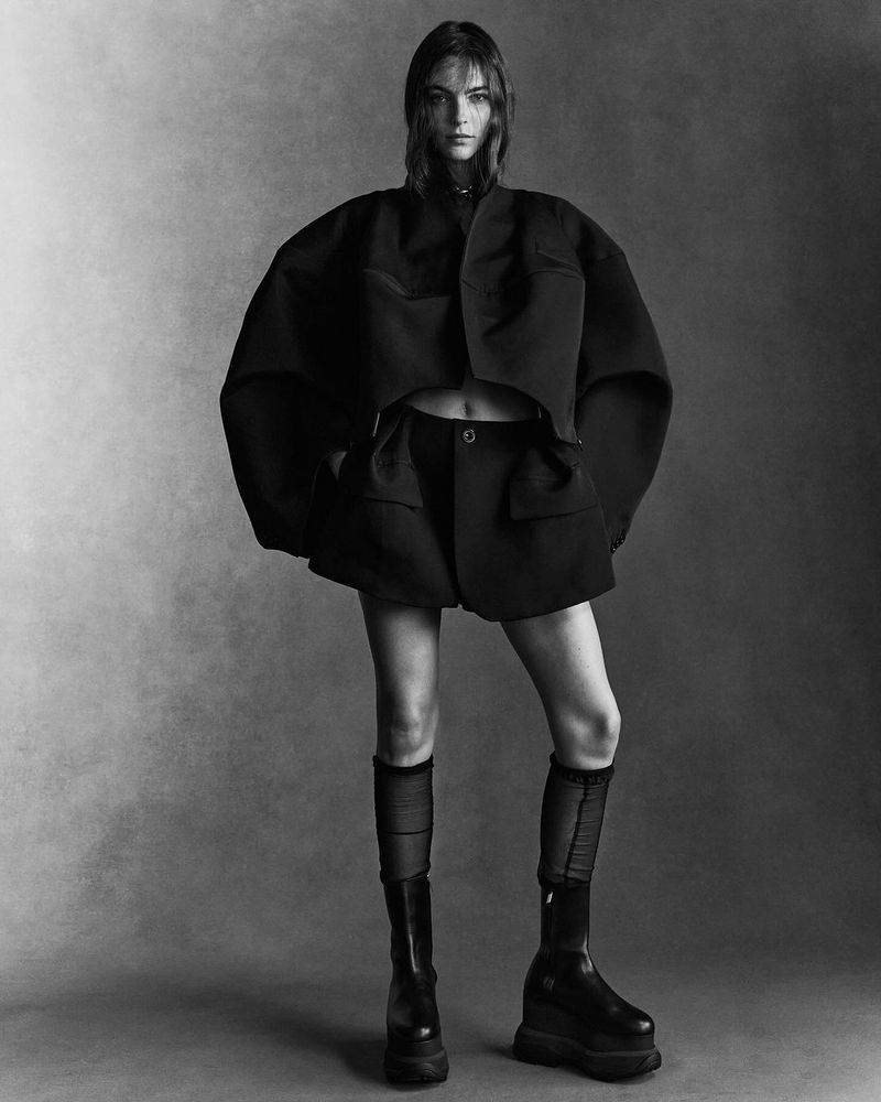 Monochrome fashion portrait of a woman posing in a black cropped jacket, black shorts, knee-high socks, and platform boots against a gray background.