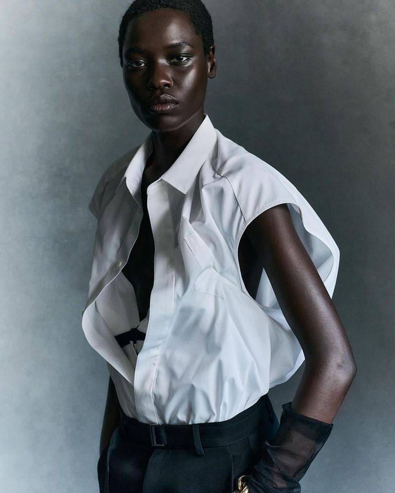 Dark-skinned woman in a white sleeveless shirt and black pants poses against a grey studio backdrop, highlighting fashion and beauty.