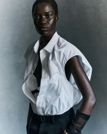 Model wearing a white structured sleeveless shirt and dark trousers against a grey studio backdrop.