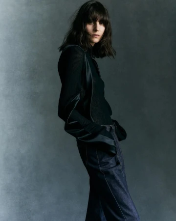 Model with dark shoulder-length hair wearing a black long-sleeve top and dark trousers against a mottled grey background