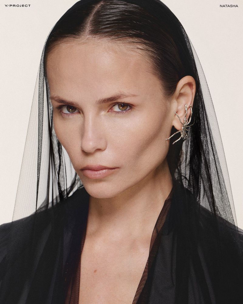 Close-up portrait of a woman with dark hair wearing a black tulle veil and a silver spider ear cuff, gazing directly at the camera.