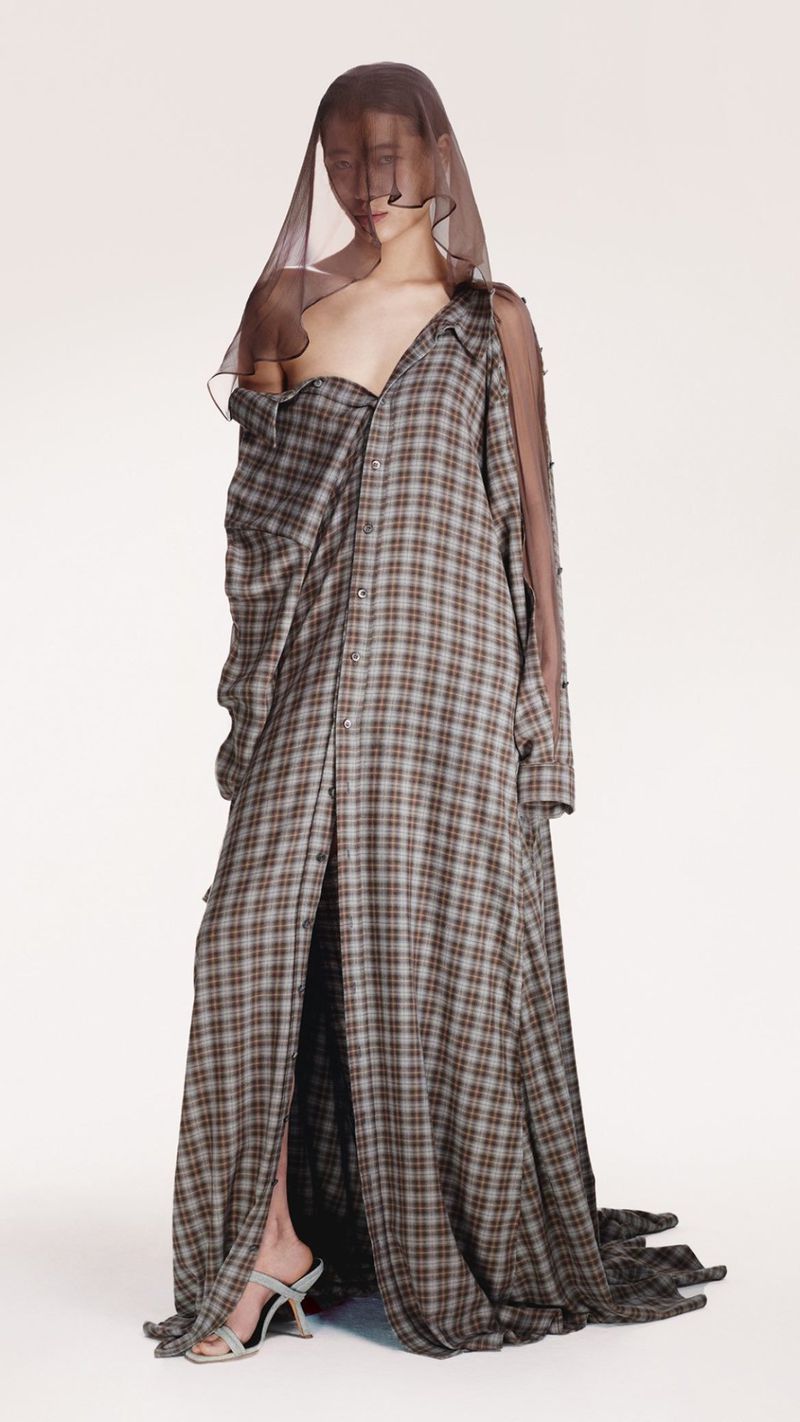 Full-length shot of a model wearing a plaid shirt dress and veil, set against a white backdrop in a fashion studio.