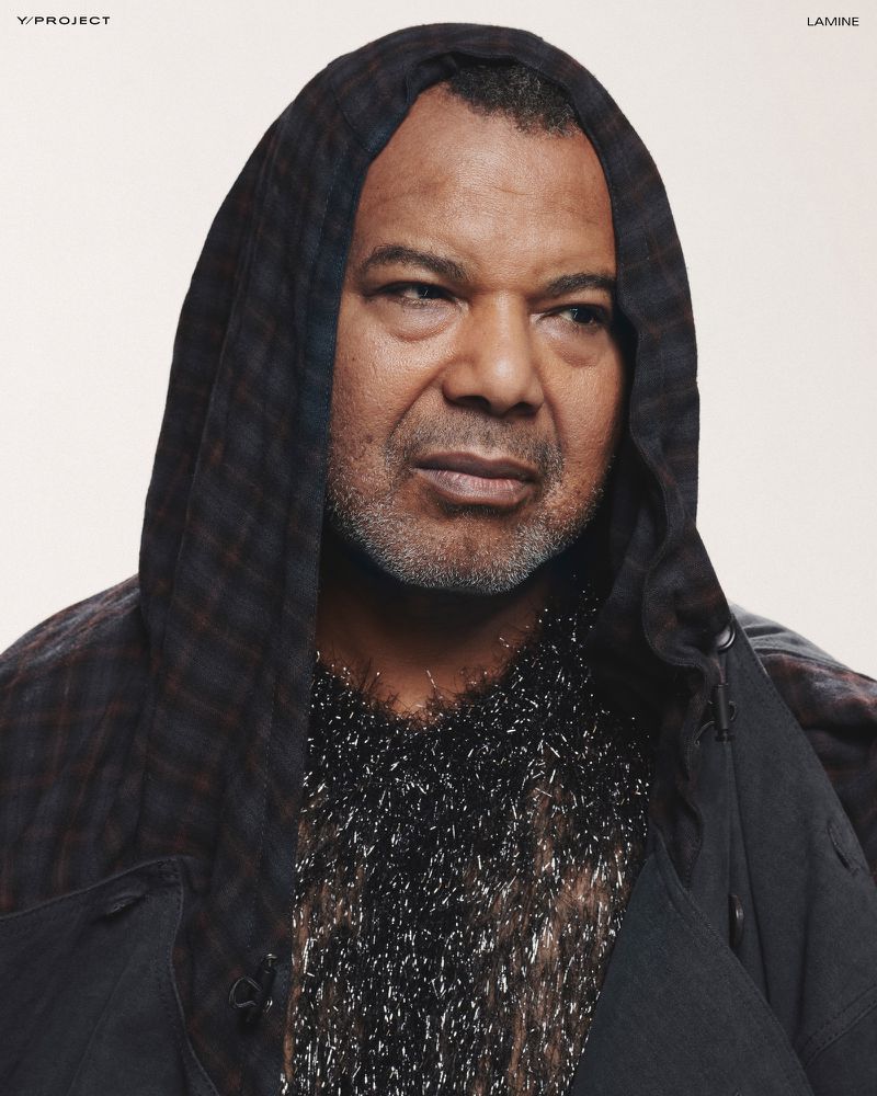 Portrait of a mature adult male wearing a hooded jacket with decorative embellishments, standing indoors against a light background.
