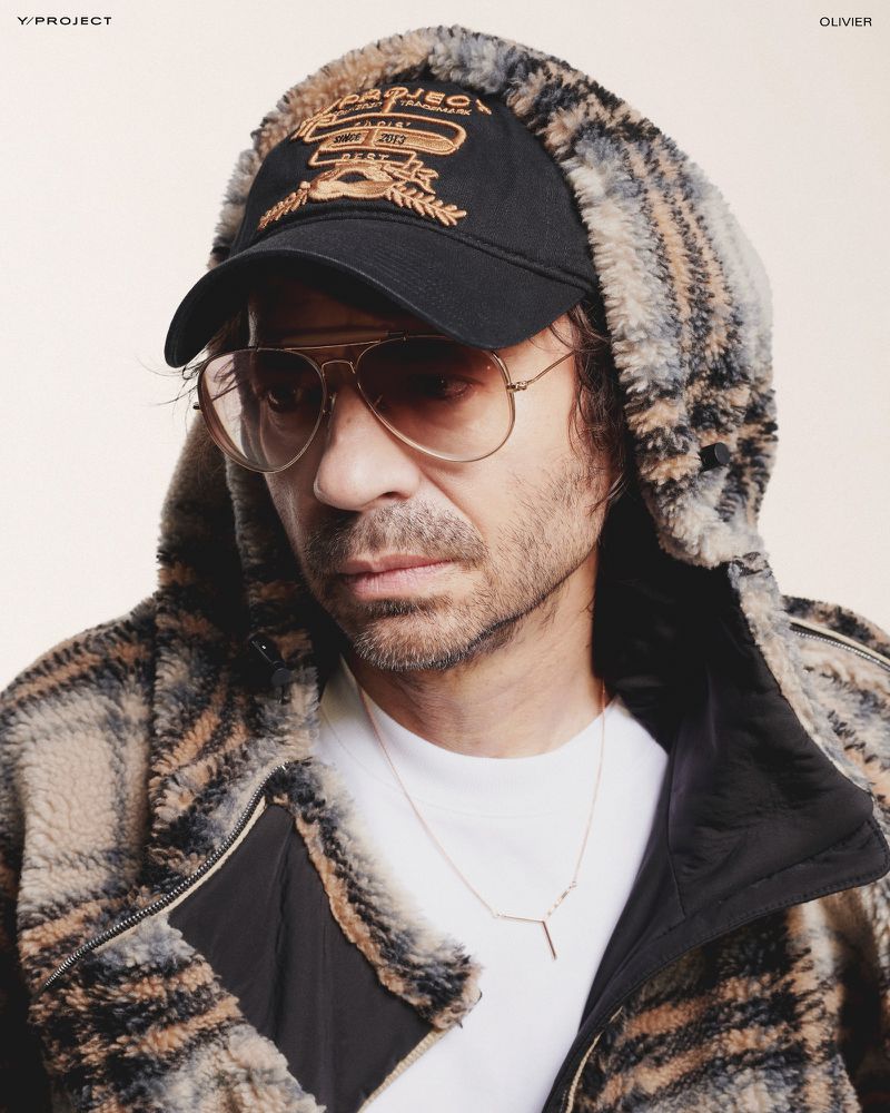 Adult male wearing a hat, sunglasses, and a hooded, patterned jacket in a close-up studio shot against a neutral background.