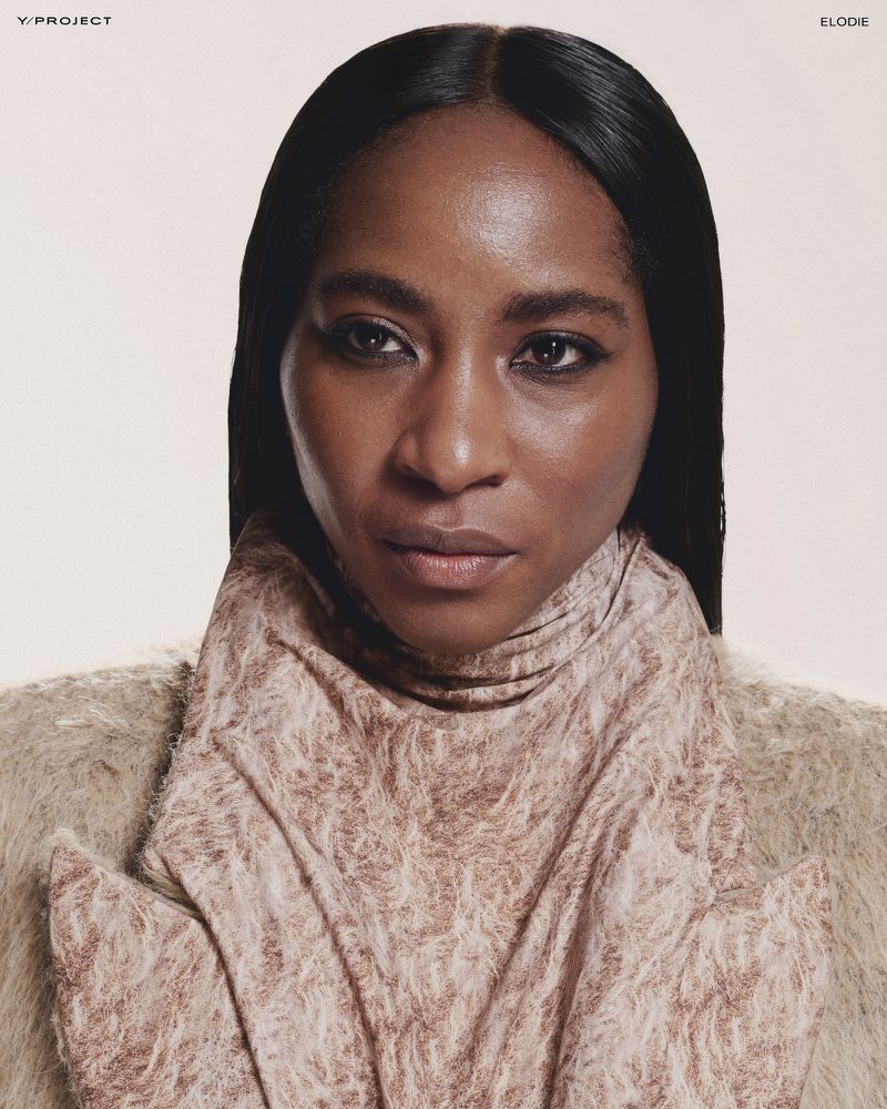 Close-up studio shot of an adult female with dark hair and skin, wearing a textured tan top. Makeup emphasizes eyes.