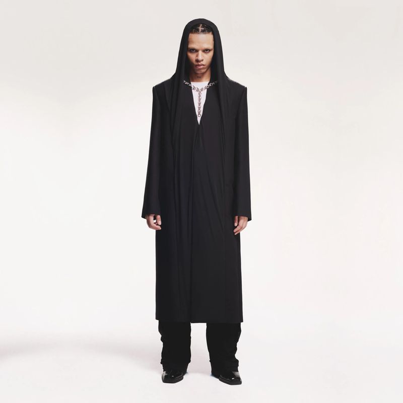 Full-length portrait of an androgynous person with dreadlocks wearing a long black hooded coat and silver chain against a white backdrop.
