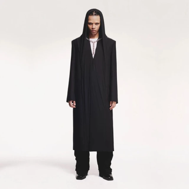 Full-length portrait of an androgynous person with dreadlocks wearing a long black hooded coat and silver chain against a white backdrop.