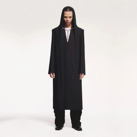Full-length portrait of an androgynous person with dreadlocks wearing a long black hooded coat and silver chain against a white backdrop.