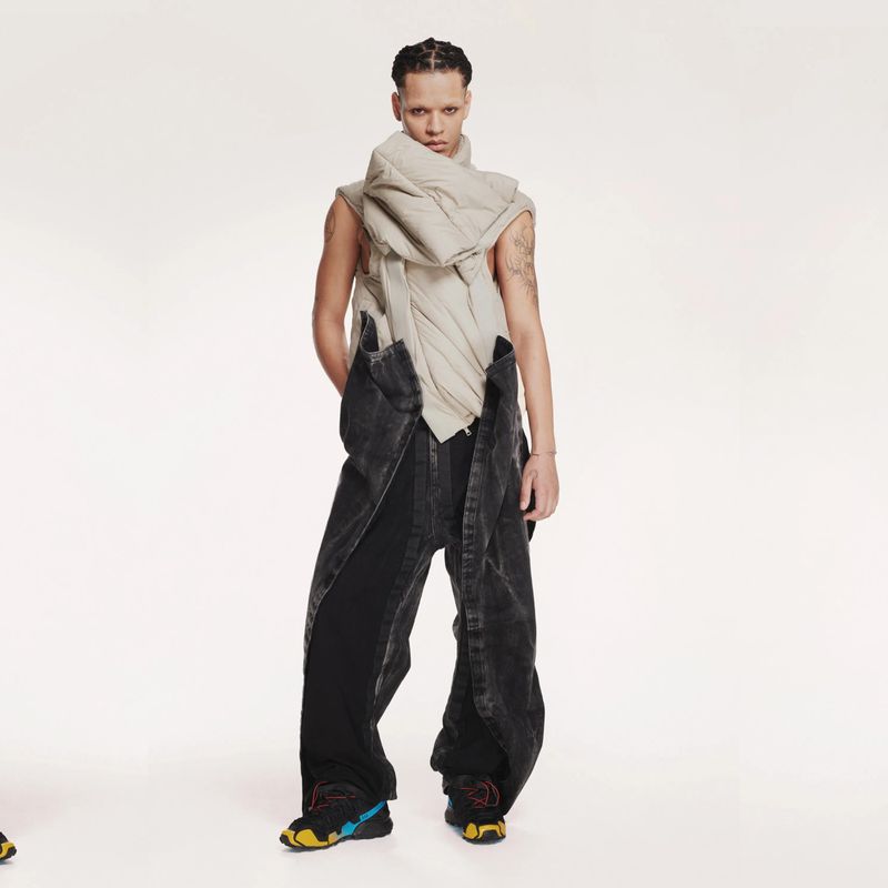 Fashionable individual poses in studio wearing puffy scarf, dark denim overalls, and athletic shoes in a full-length shot with bright lighting.