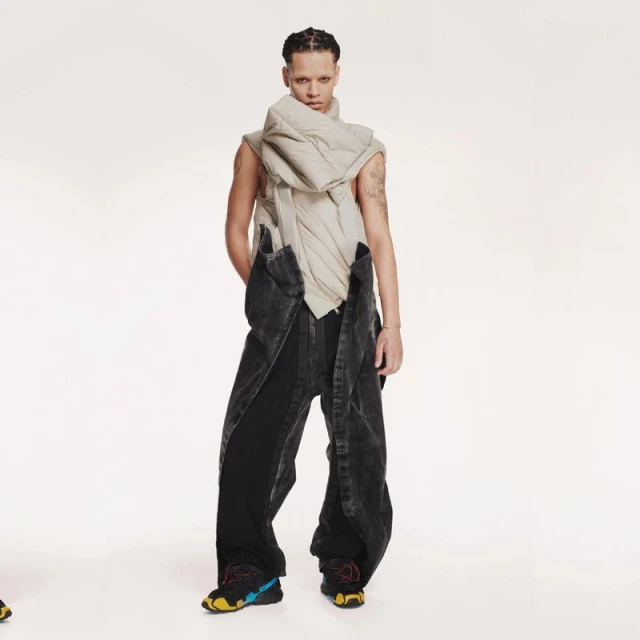 Fashionable individual poses in studio wearing puffy scarf, dark denim overalls, and athletic shoes in a full-length shot with bright lighting.
