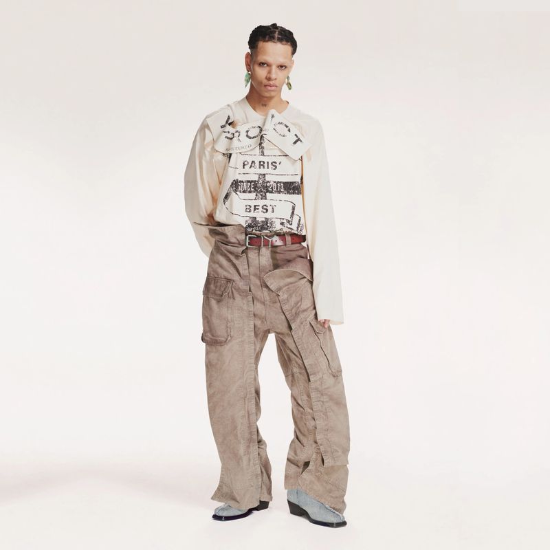 Full shot of person in tan long-sleeve shirt with graphic print and wide-leg cargo pants against a white backdrop in a studio.