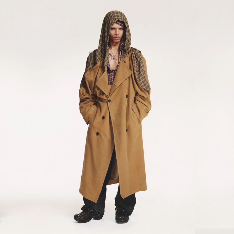 A person in a long, light-brown trench coat and patterned hood poses in front of a white background in a fashion-style photograph.