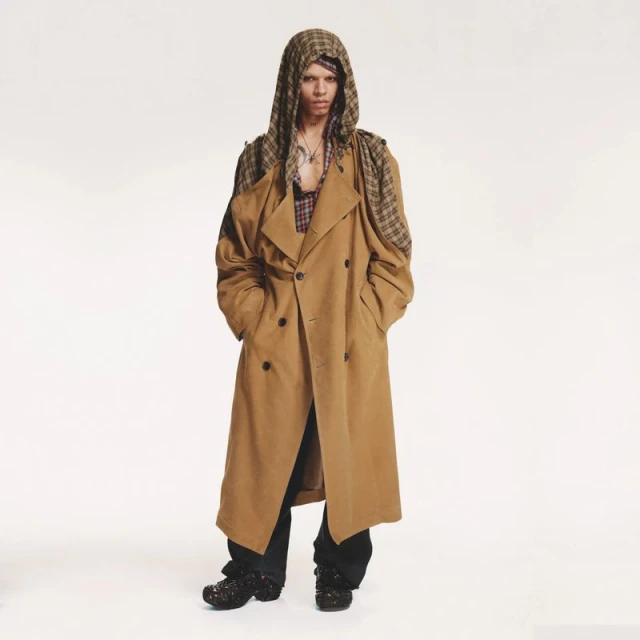 A person in a long, light-brown trench coat and patterned hood poses in front of a white background in a fashion-style photograph.