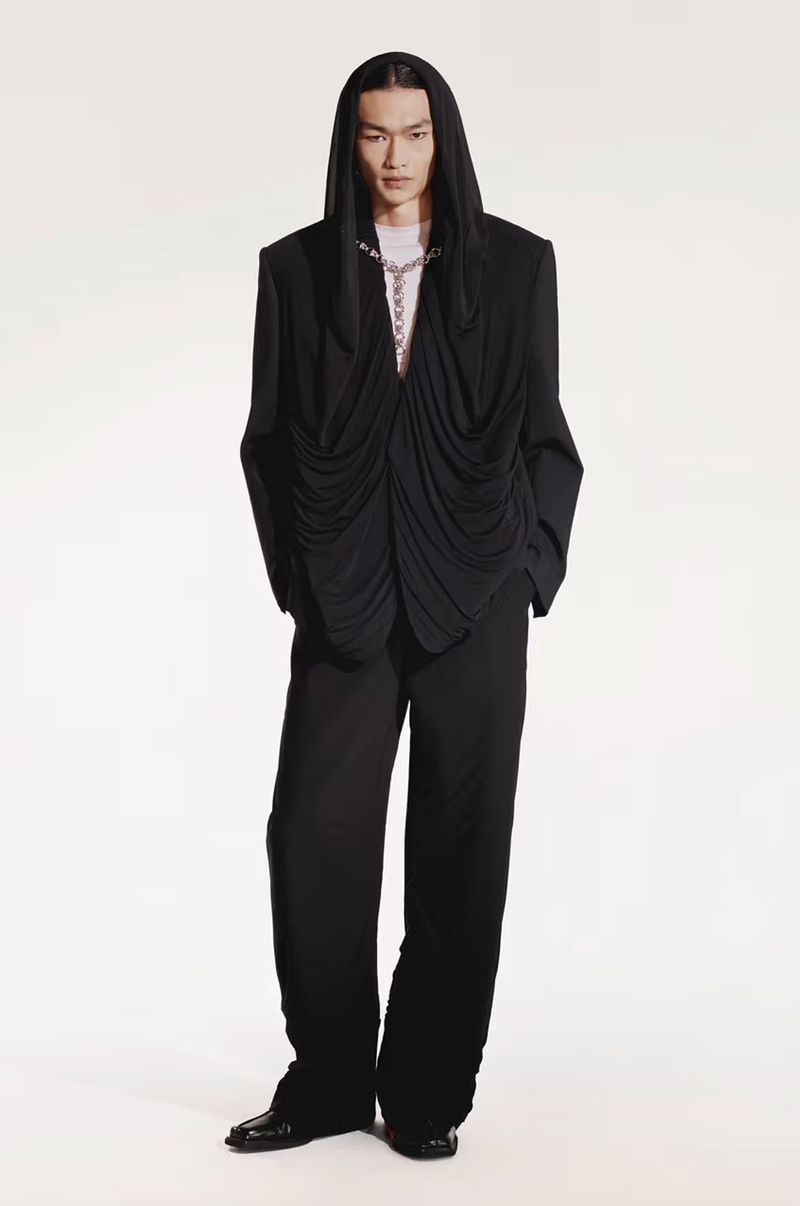 Asian male fashion model in black hooded draped jacket and wide-leg pants against white background in studio fashion shot.